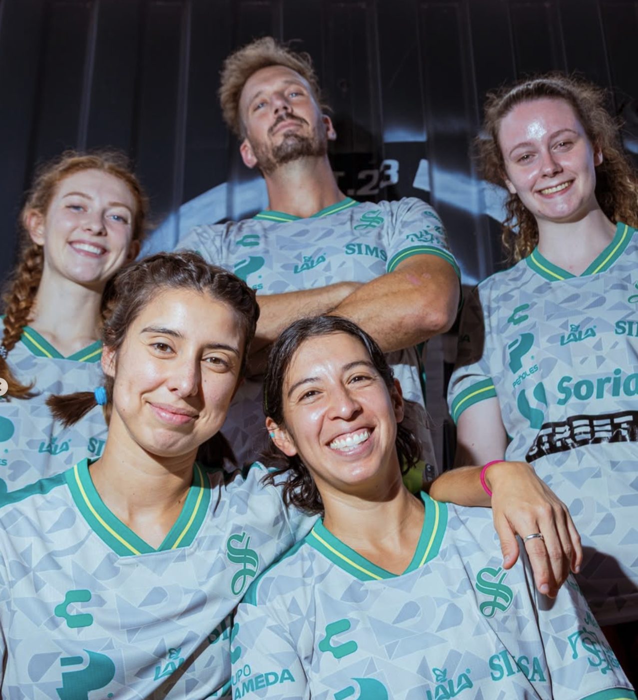 Five teammates in white and green jerseys together in front of the Barrio wall